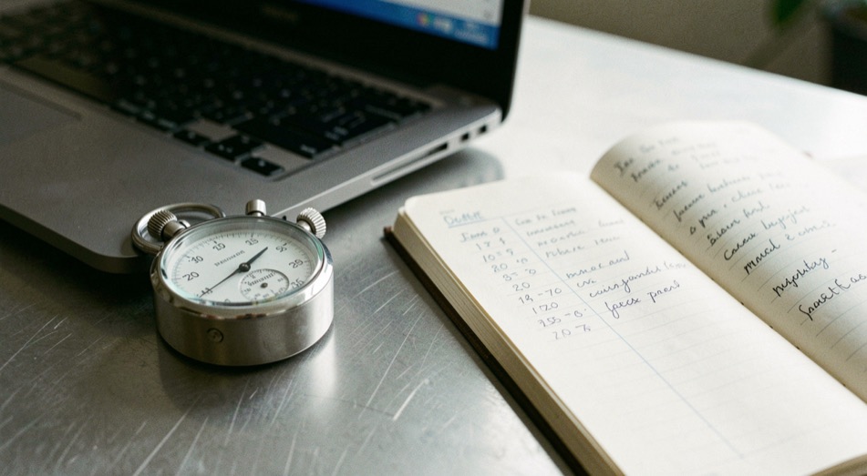 Un chronomètre mécanique posé à côté d'un ordinateur portable sur un plan de travail en métal brossé, avec un carnet ouvert sur une colonne de mesures manuscrites