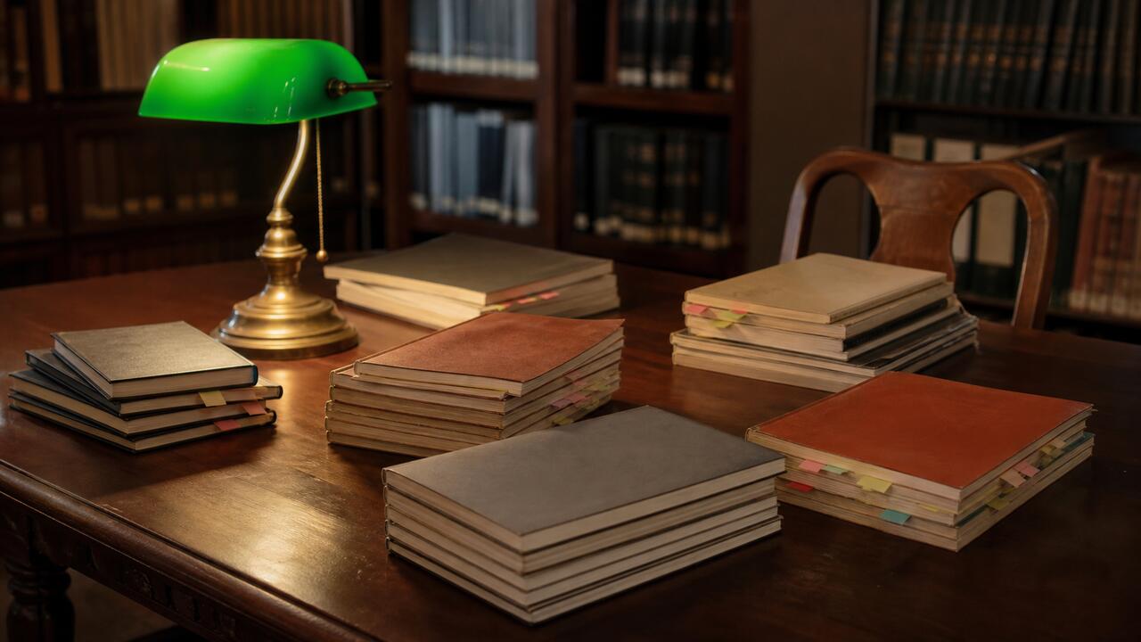 Une table de bibliothèque en bois sombre couverte de revues techniques empilées, marque-pages colorés dépassant des tranches, lampe à abat-jour vert allumée