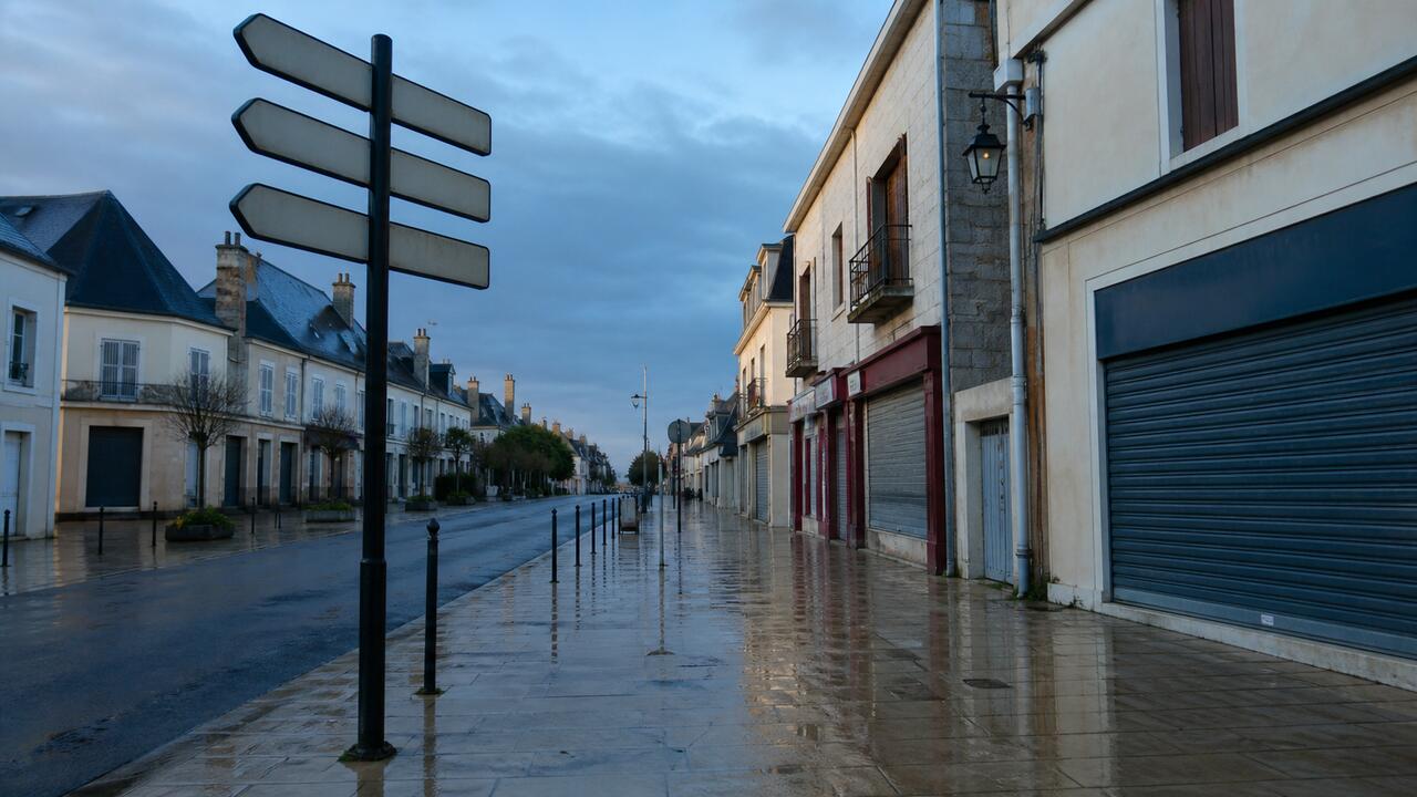 Une rue commerçante de centre-ville en début de matinée, une devanture aux stores encore baissés, un panneau indicateur et des pavés humides après la pluie.